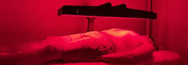 Person lying on a treatment table beneath a red light therapy panel in a red-lit room.
