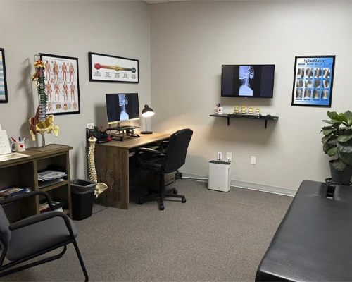Consultation room with anatomical posters, spine model, monitor, shelving, chairs, and a stool.