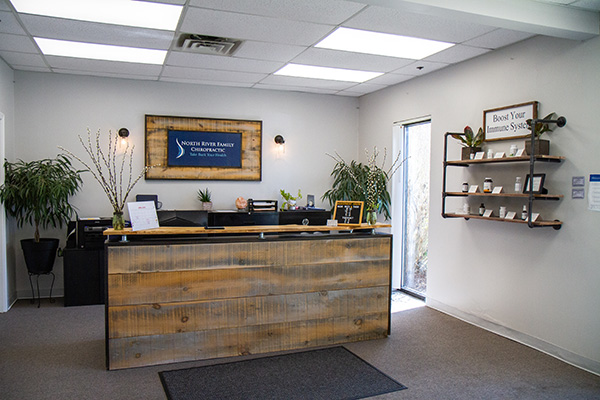 Reception area with a wood-front desk, shelving, plants, and chiropractic office signage on the wall.