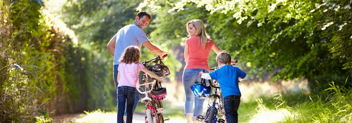 Family walking with bicycles along a tree-lined path in an outdoor setting.